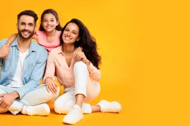 Une jeune famille, le père, la mère et une petite fille, souriants, assis par terre, sur fond jaune.